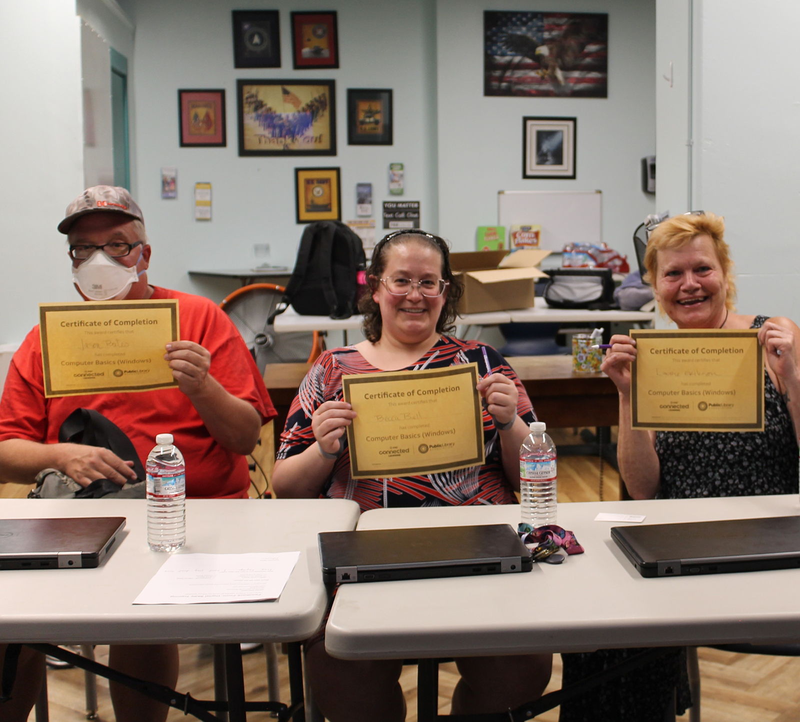 Four attendees of a computer basics class hold up their ccompletion certificate, along with their free laptops
