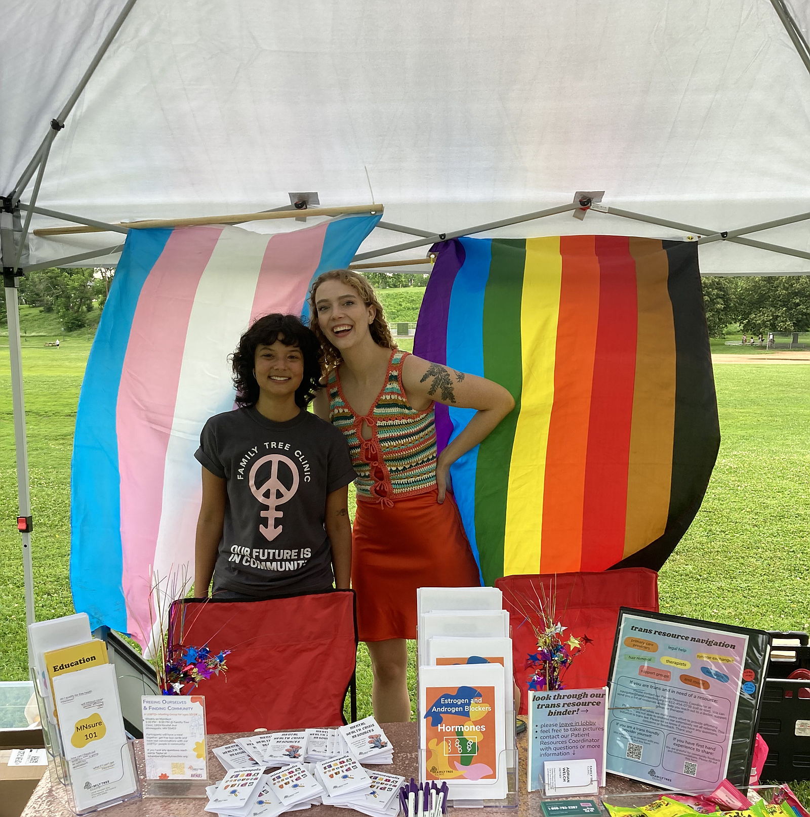 Two smiling volunteers at the Family Tree Clinic Pride booth. Pride flags are waving in the background.