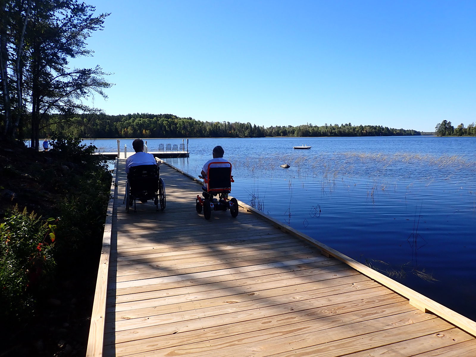 Two women using wheelchairs rolling side by side on an accessible boardwalk over a lake on a clear sunny day.