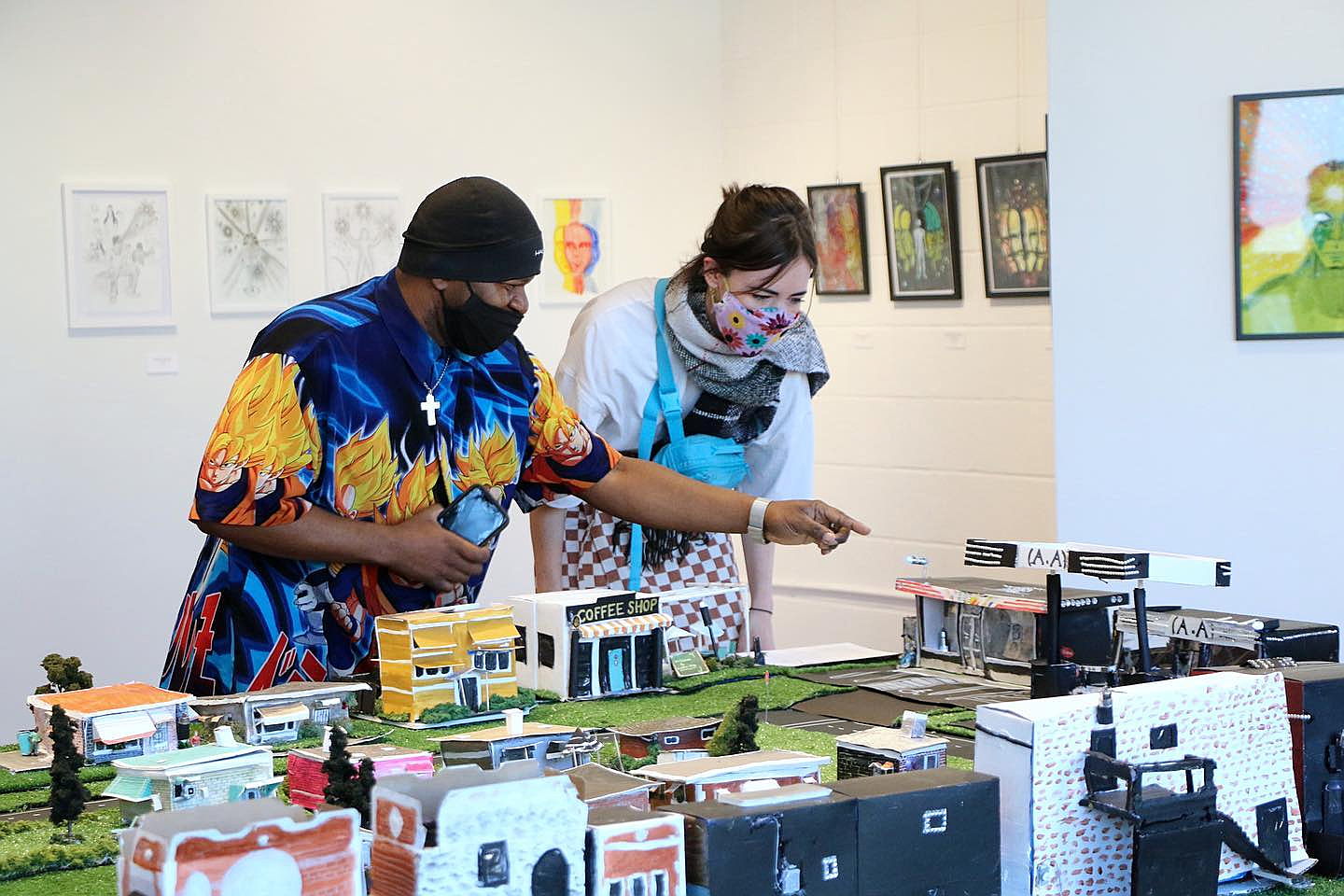 A man showing a woman his miniature city art piece. It sits a top a large table.