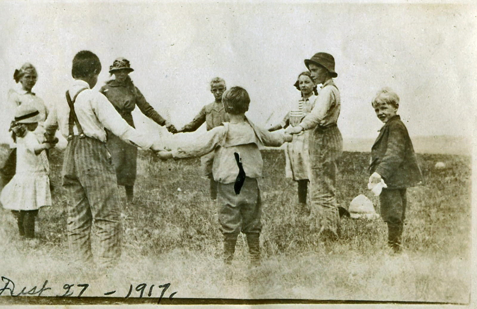 vintage 1917 photo of children holding hands in a grassy field. one boy on the right side of the image looks at the camera