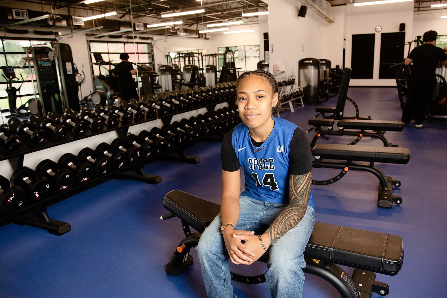 Image of a student wearing a blue jersey with braided black hair sitting on a weight bench in a weight room. 