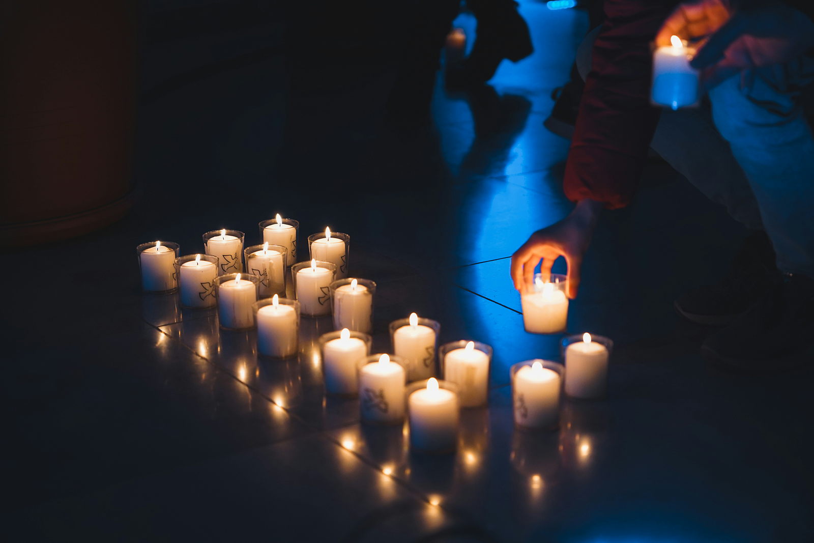 Rows of votive candles in clear glasses providing the only illumnation in the scene, set on a shiny black tiled floor in a dark room as more hands are lining up to add more lit candles to the array