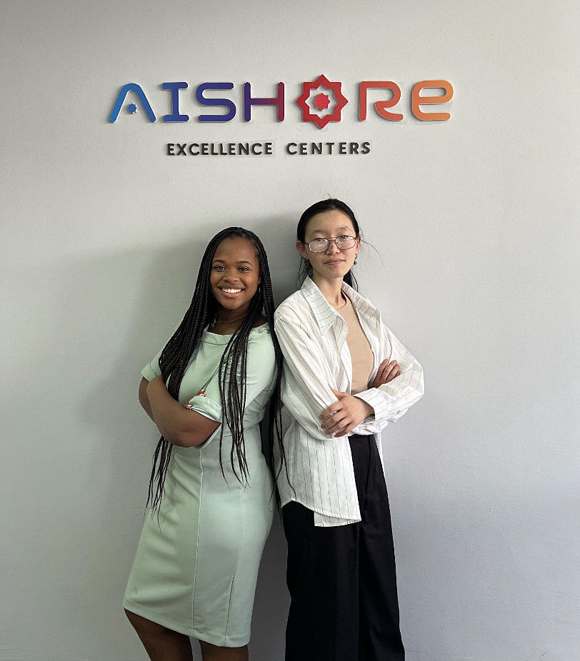 Two women stand back to back in front of sign for Aishore Excellence Centers