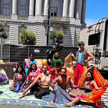 A group of dancers sits on a large piece of embroidered cloth in bright sunlight. They are dressed in performance attire and holding multicolored scarves.