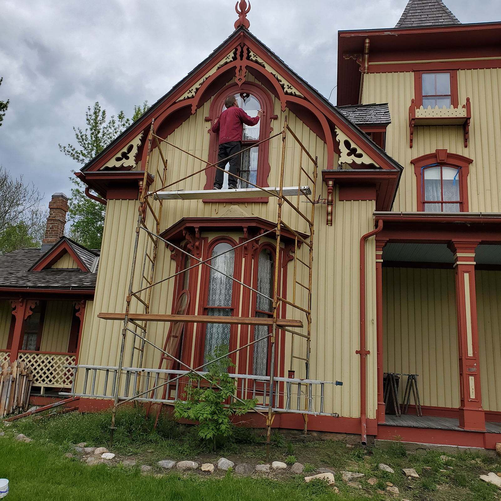 Yellow and red house with scaffolding in front of it with a man repairing a second story window.