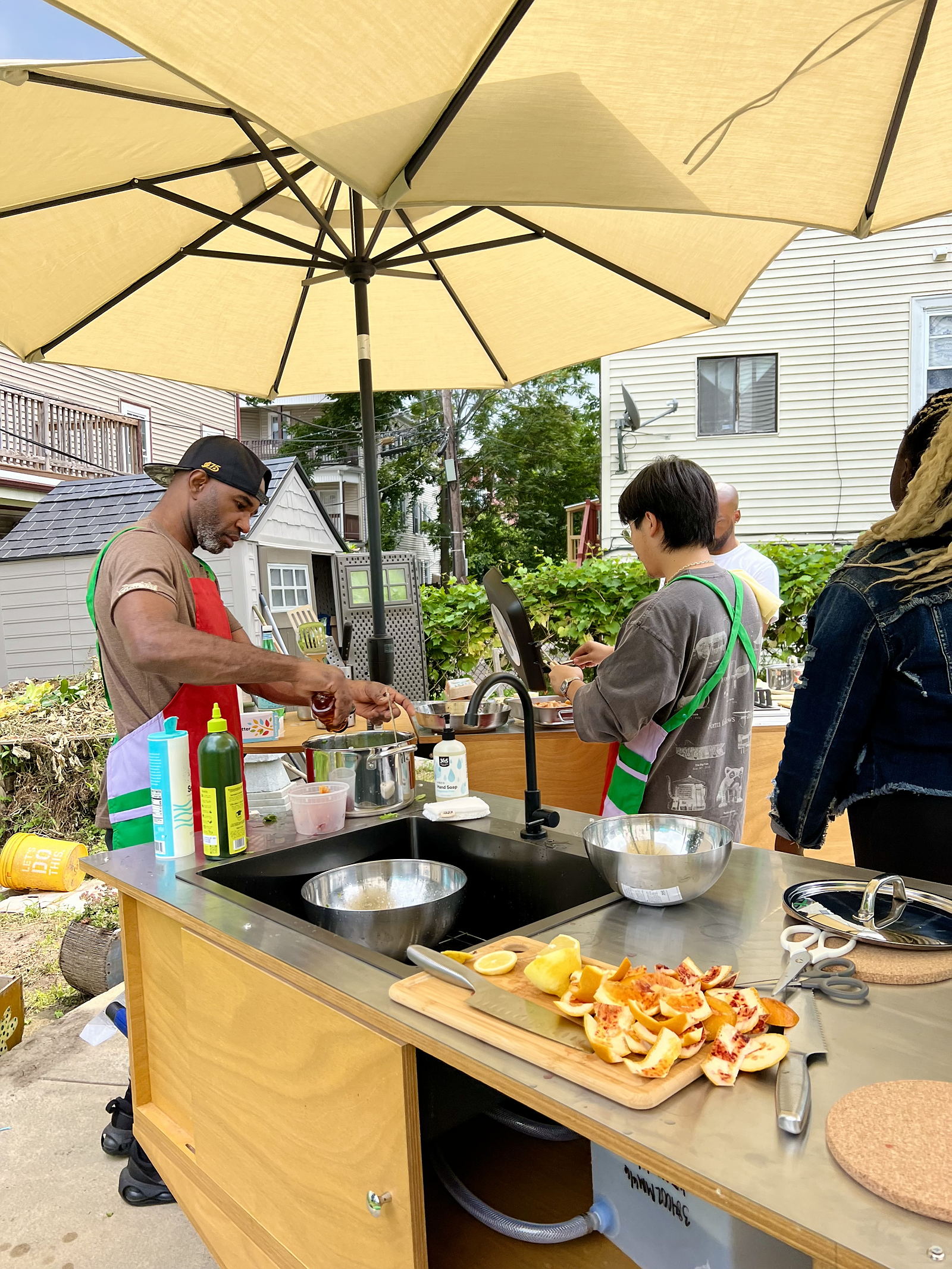 Two men wearing vibrant red aprons, stand in an outdoor kitchen, surrounded by a green garden, and cook on a stovetop.