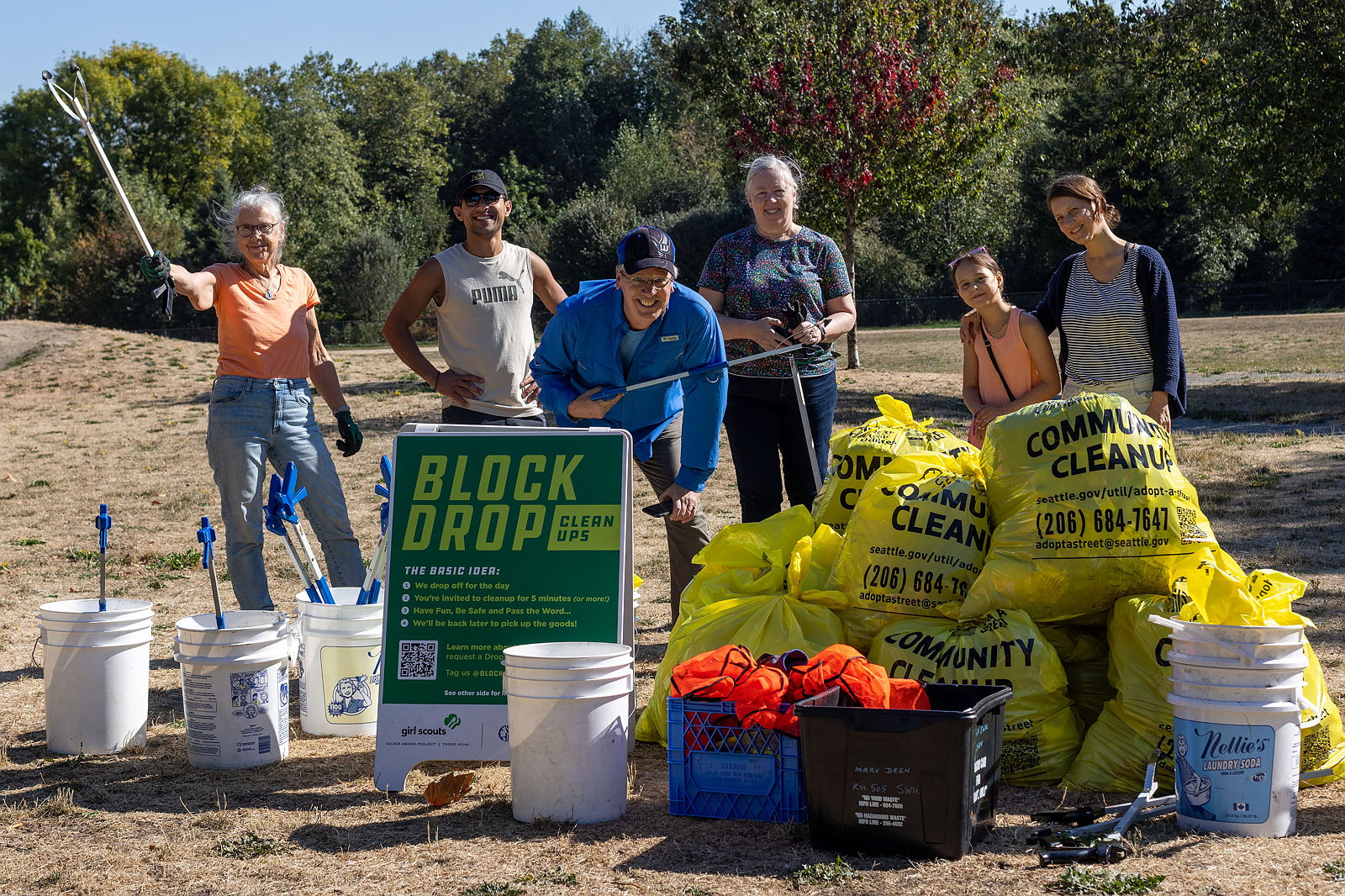 Six people stand smiling with tools in hand behind a sign that reads Block Drop and yellow cleanup bags.