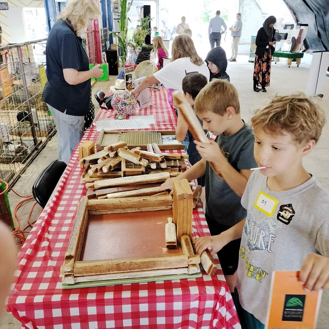Children participate in a tabletop log cabin building activity at a table covered with a red and white checkered tablecloth, while adults assist in the background.