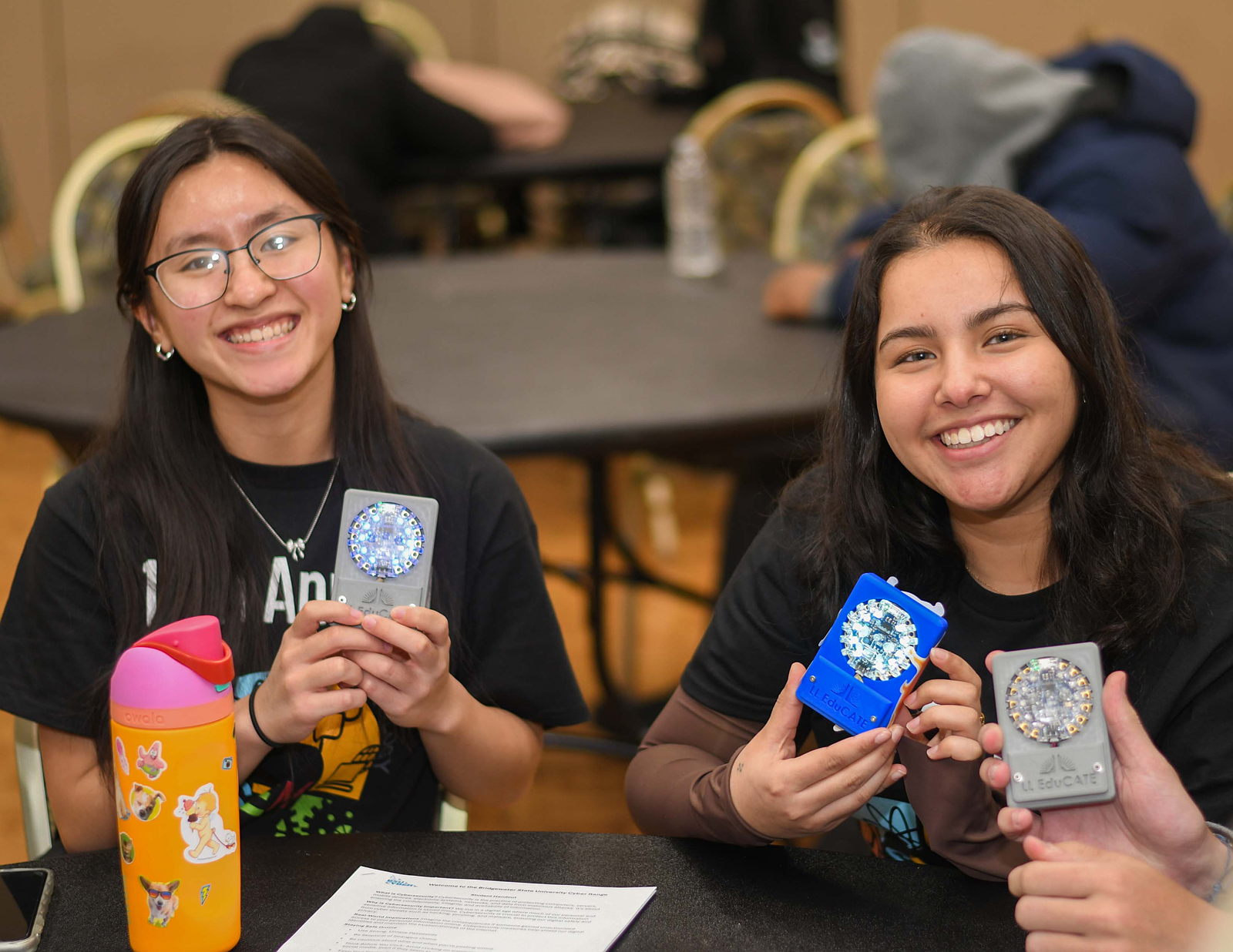 Two smiling female students seated at a worktable show off their custom LL EduCATE Development Boards.