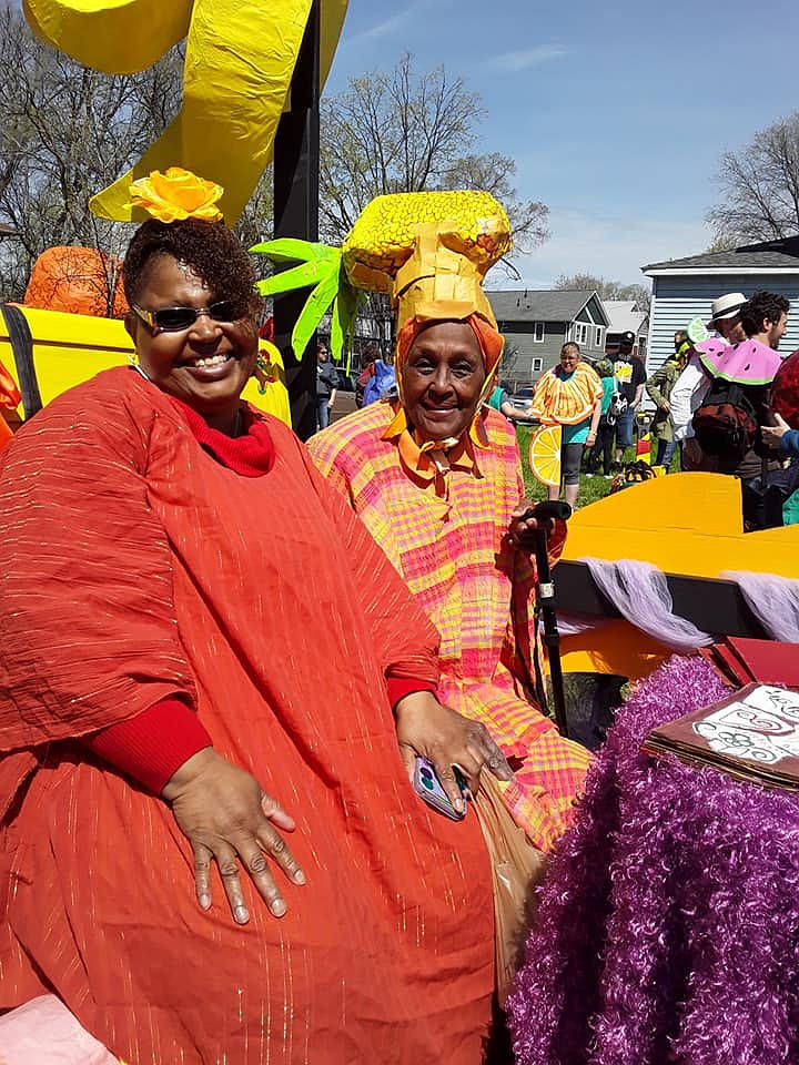 Two zAmya Troupe Members sitting on a float in the May Day Parade. They are dressed in bright reds and yellows.