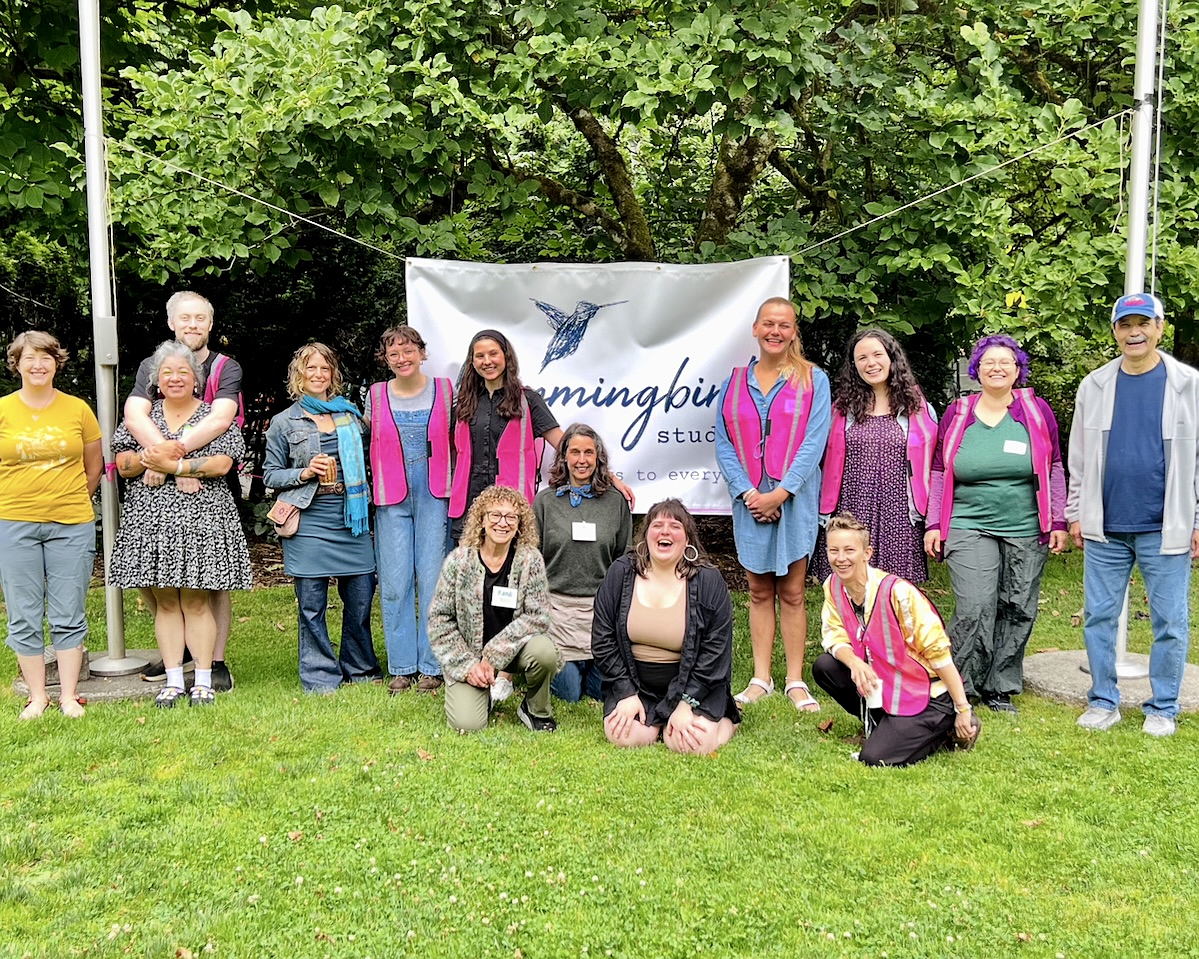 hummingbird studio celebrated its 10th birthday at Squaxin Park, and here volunteers and event helpers gather on the grass in front of the studio banner in a wide group shot. Several are wearing hot pink safety vests. 