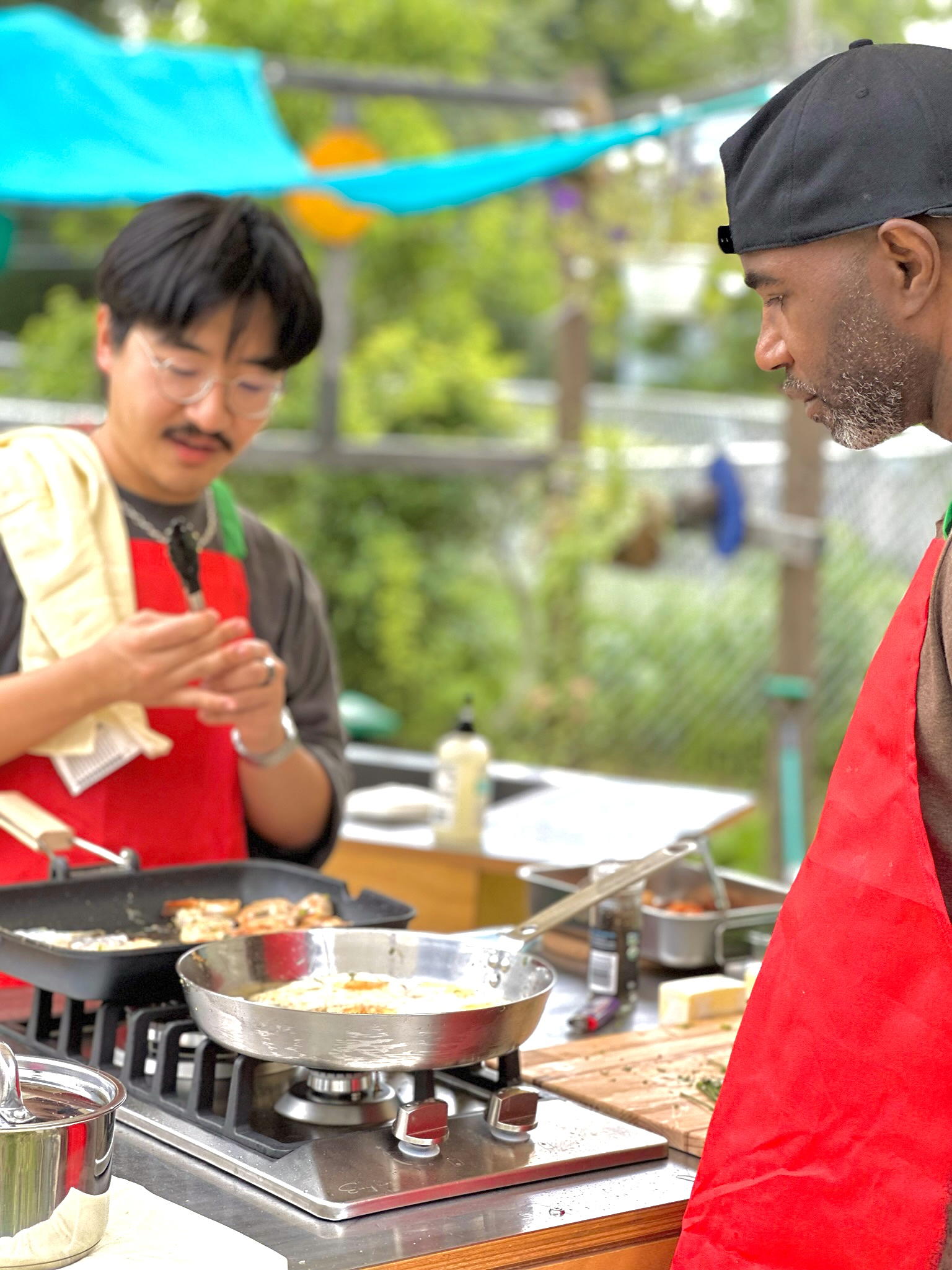Two men wearing vibrant red aprons, stand in an outdoor kitchen, surrounded by a green garden, and cook on a stovetop.