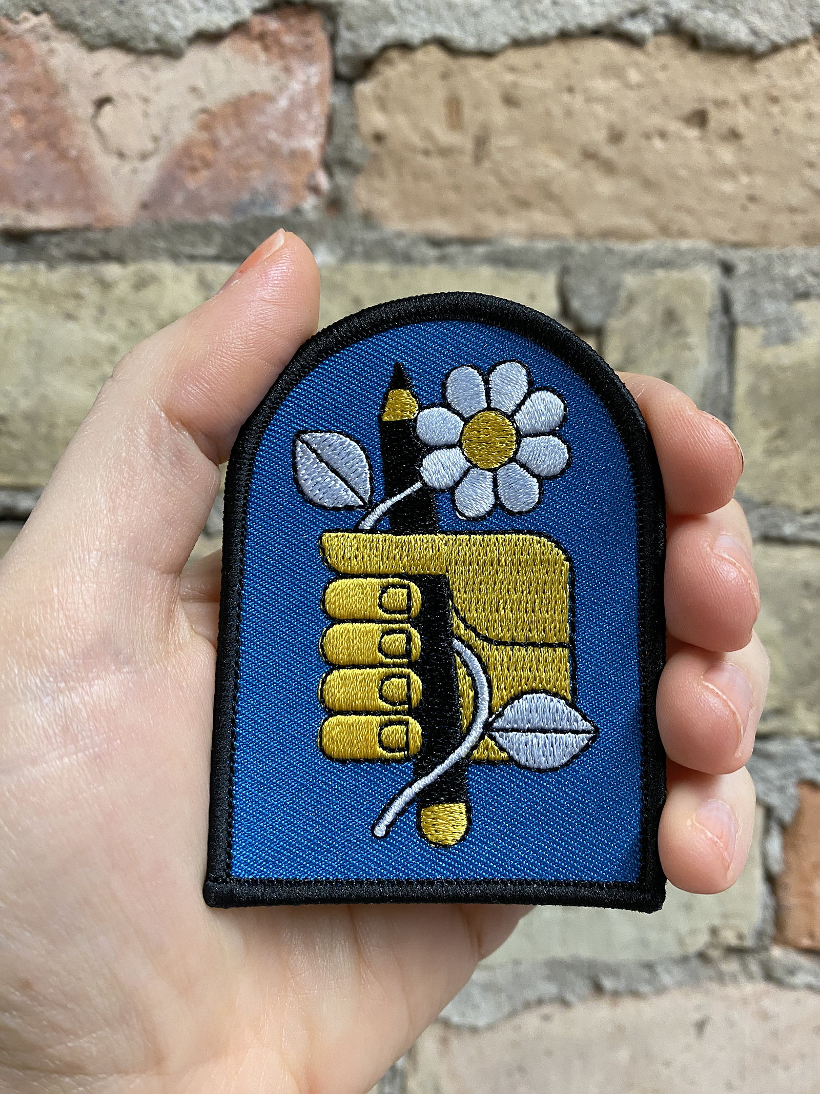 A patch of a yellow hand holding a black pencil and white flower, stitched in a bold graphic style. There is a blue background and black trim around the edge of the patch.