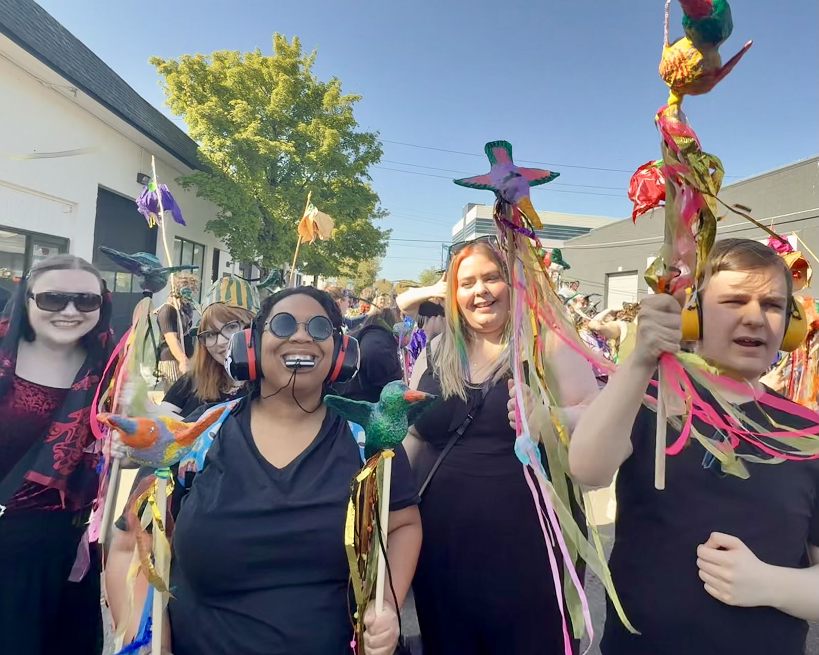 Artists. all dressed in black, celebrate The Procession of the Species parade holding brightly painted paper clay hummingbirds on wooden sticks decorated with multi-colored ribbon. 