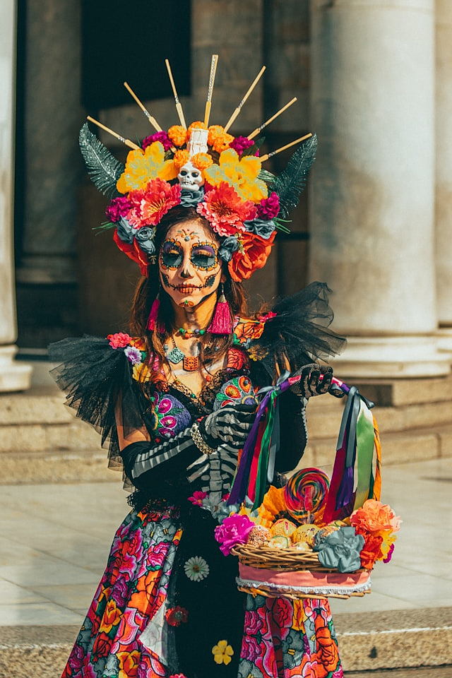 A photo of a woman in a brightly colored dress decorated with Day of the Dead symbols in Mexico City, Mexico and wearing makeup invoking the image of the dead holding a basket of flowers on a bright day in a street scene.