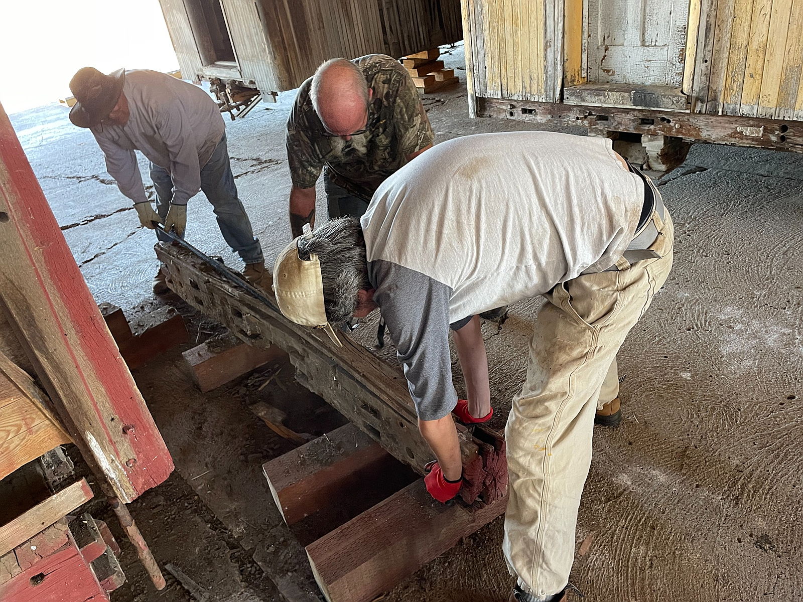 Three volunteers lift a deteriorated end sill from Boxcar No. 518, exposing the supporting timbers beneath it.