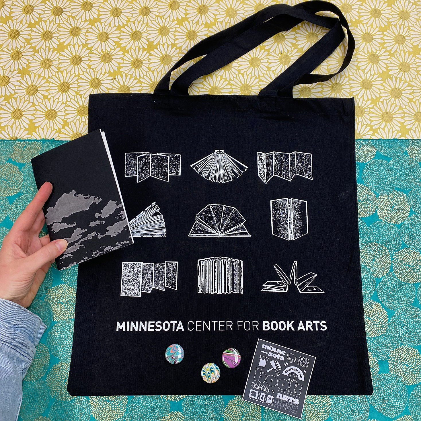 A black tote bag with a print in white ink. The print features nine illustrations of books, with text reading, "Minnesota Center for Book Arts" at the bottom.