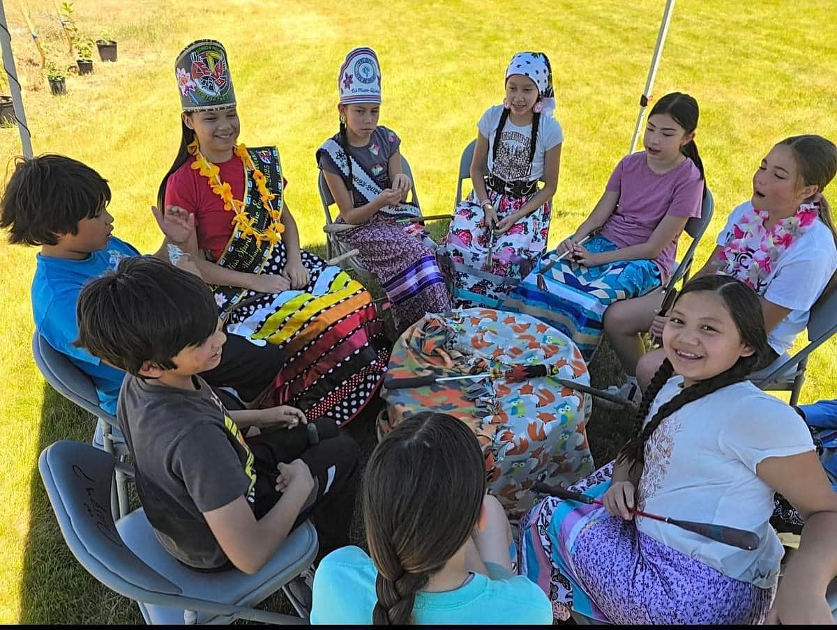 Spokane Salish School- Potlatch Grantee. Kids sitting round a round drum in ribbon skirts, regalia, and smiling