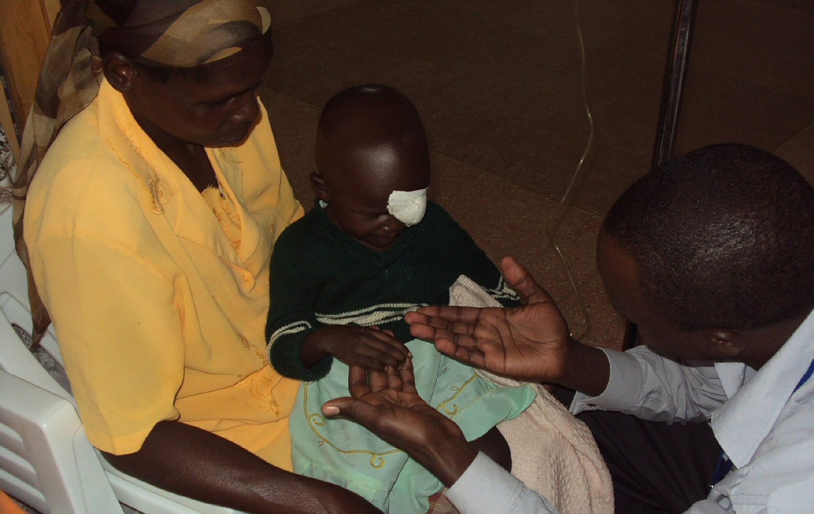 A young girl with a bandage over her eye sits on her mother's lap. A medical profesisonal holds out his hands to her, allowing her to make the first physical contact before a medical procedure.