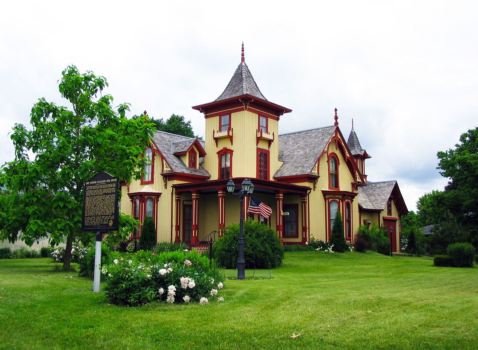 Yellow and red house pictured on a green lawn with various trees in the background and a peony bush in the foreground. 