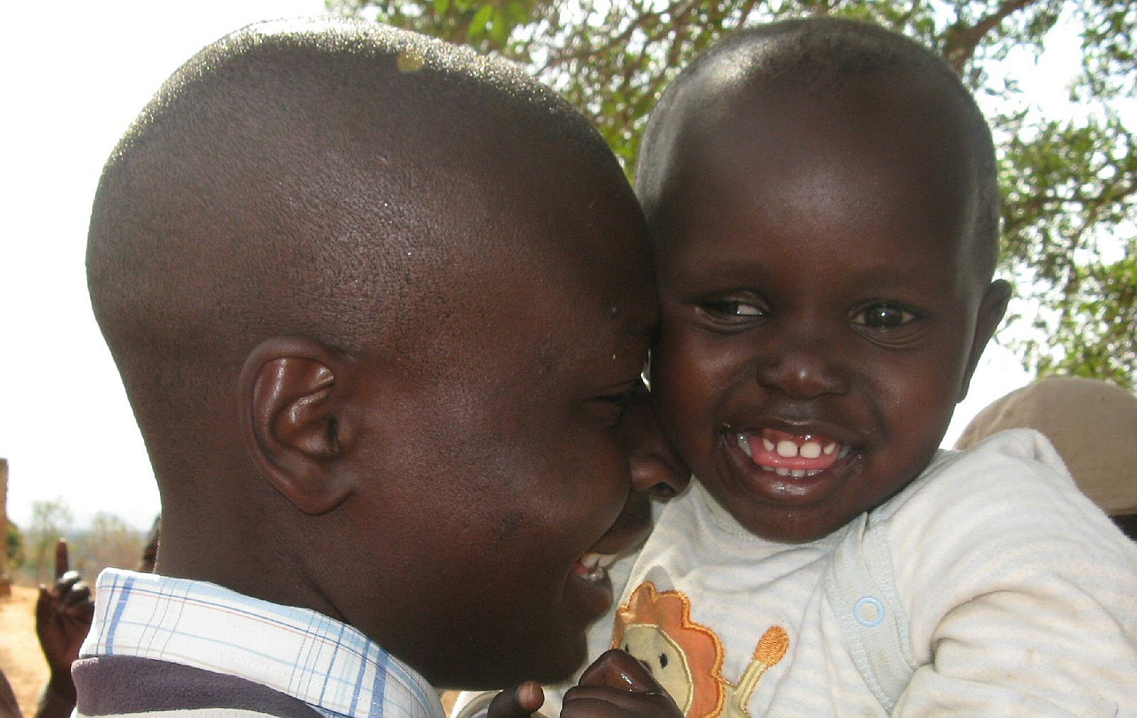 A young girl rests in her father's arms as they smile at one another. One of her eyes has a white pupil glow, and the other has an outward turned squint. The background is a rural African village.