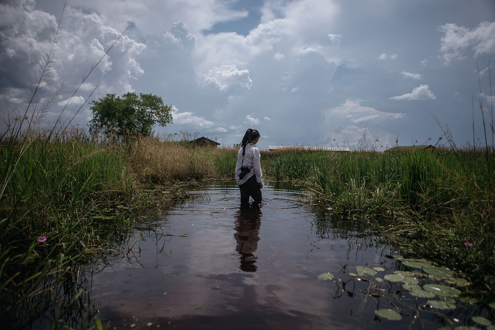 An Asian woman in a white shirt and ponytail, with a camera strapped to her back,wades through knee-deep water