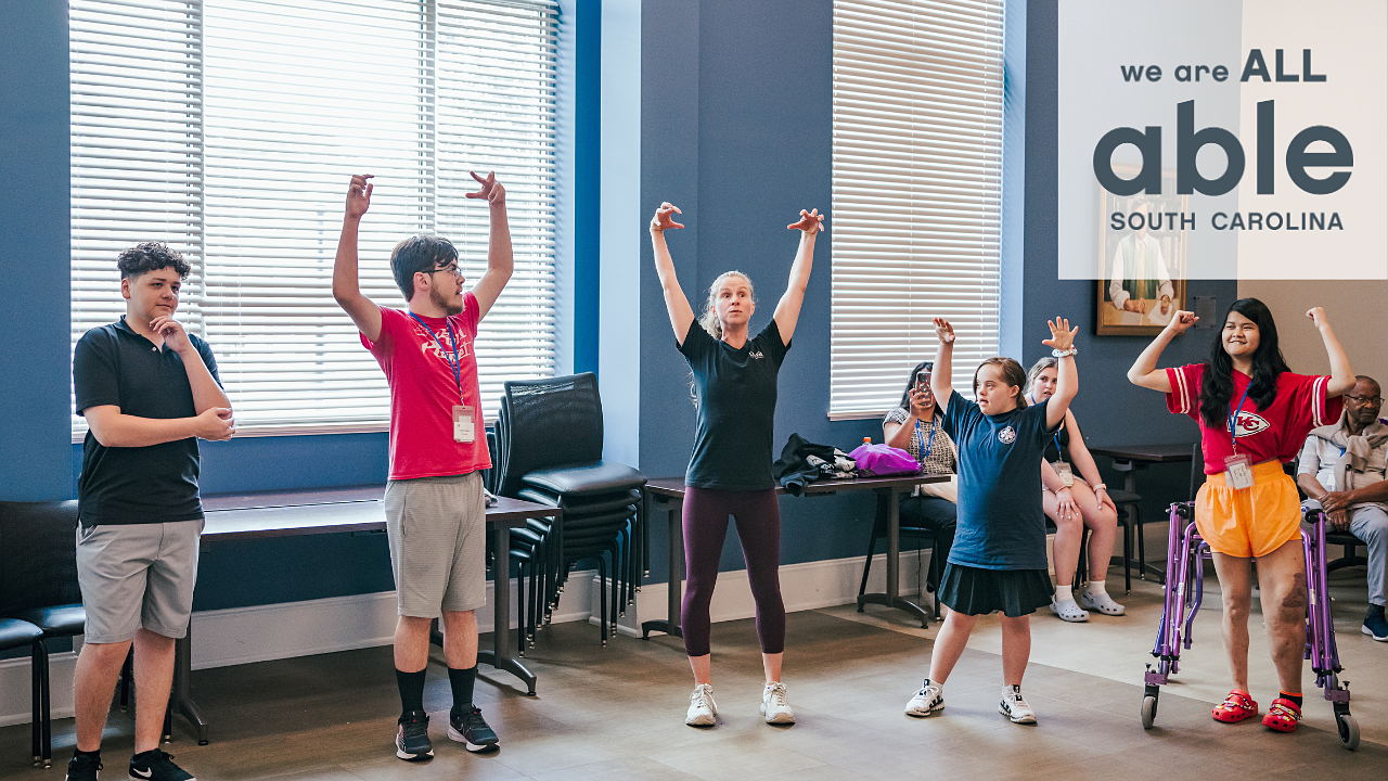 A group of four young disabled people, two young men and two ladies, follow the instructor, in the center, stretching their arms up high to the sky. One of the young men ponders instead. 