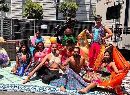 A group of dancers sits on a large piece of embroidered cloth in bright sunlight. They are dressed in performance attire and holding multicolored scarves.