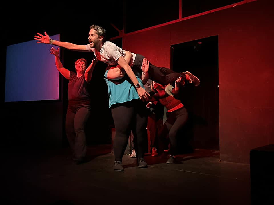An improviser being lifted by 3 others and reaching toward the audience on a colorfully lit stage.