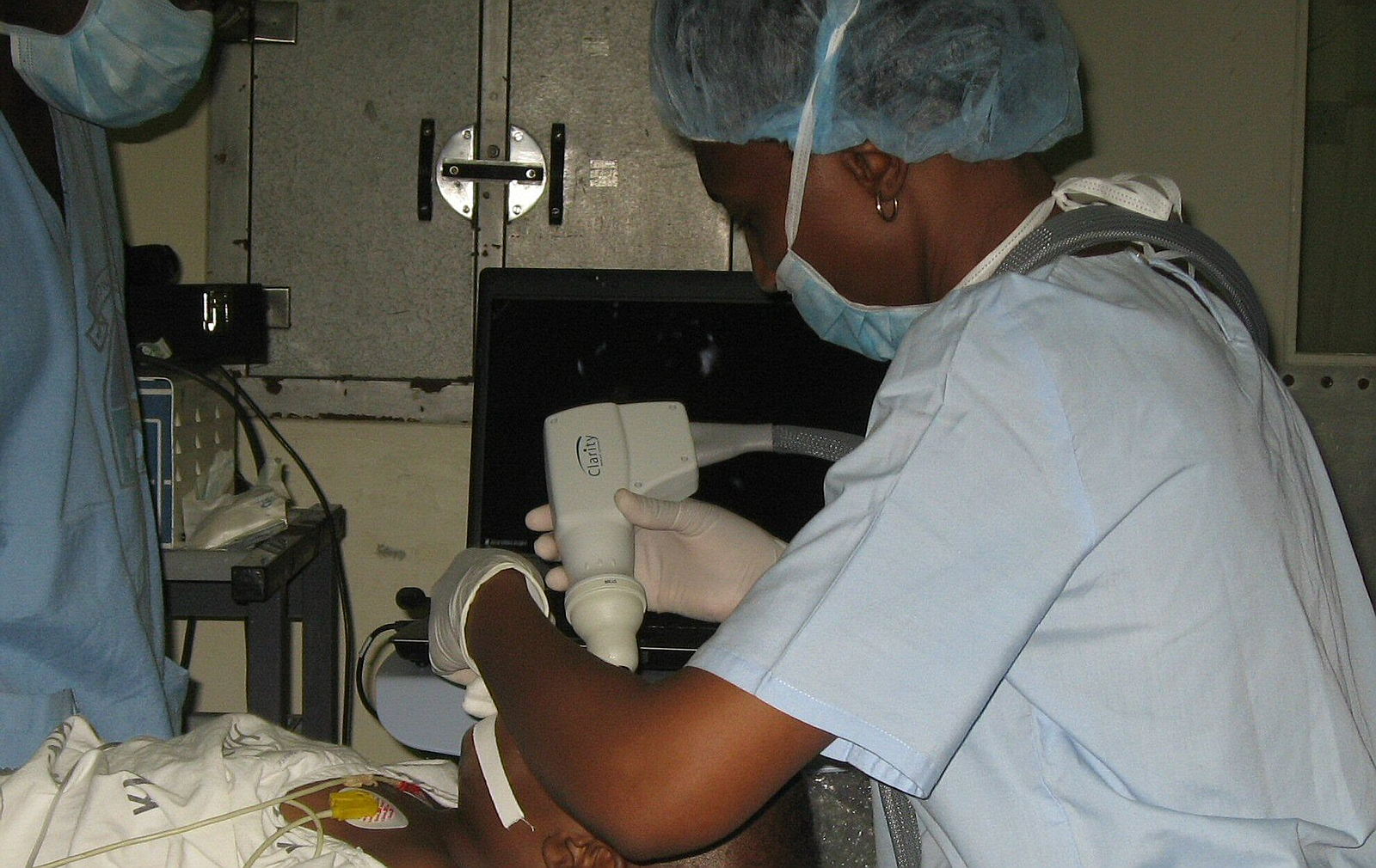 A doctor wearing scrubs, surgical cap, gloves and mask carries out an eye exam on an anaesthetised toddler. She is holding a retcam digital camera probe, and a computer sits to her side, projecting high resolution images onto the screen.