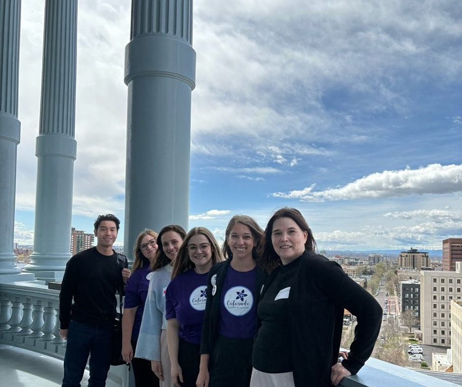 Image shows a photograph of six of the Immunize Colorado staff team standing outside the Colorado State Capitol dome observation area, with the Denver cityscape in the background.