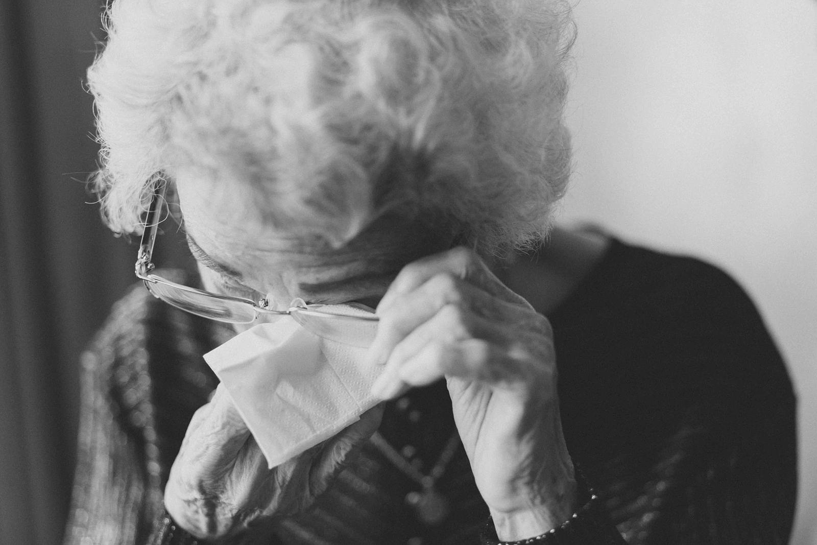 A black and white close-up photo of an older woman with silver hair, head bowed, holding her eyeglasses away from her face with her left hand allowing her to to dab the tears from her eyes with right hand