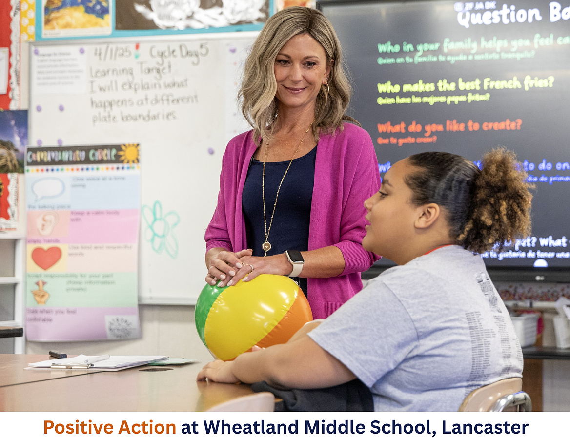 In a middle-school classroom, a teacher stands over a student desk. The student is sharing, and the teacher listens with care and concern. 